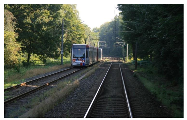 Et letbanekøretøj kører på et dobbeltsporet tracé omgivet af træer i et skovområde. Sporet er tydeligt adskilt fra vejtrafik, og letbanen reguleres med signaler, som ved en regulær jernbane.