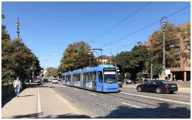 En blå letbane kører på en vej, hvor den deler tracé med biler og andre trafikanter. Letbanesporene er ikke adskilt fra resten af vejtrafikken, hvilket gør det muligt for fodgængere og køretøjer at krydse sporet frit.